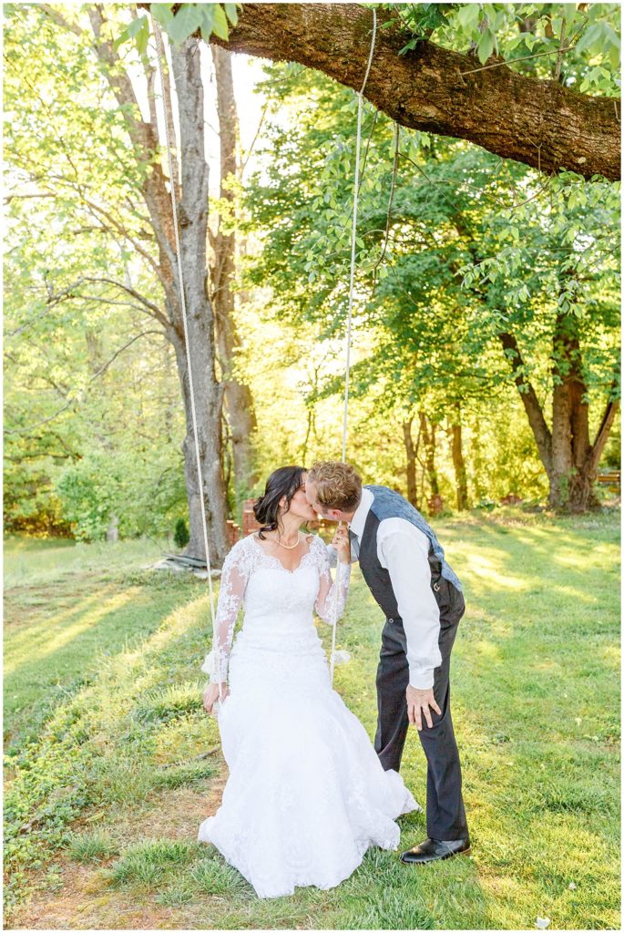 The bride sits on the swing under a tree and kisses her groom at New Beginnings Historic Farm outside Asheville, NC.