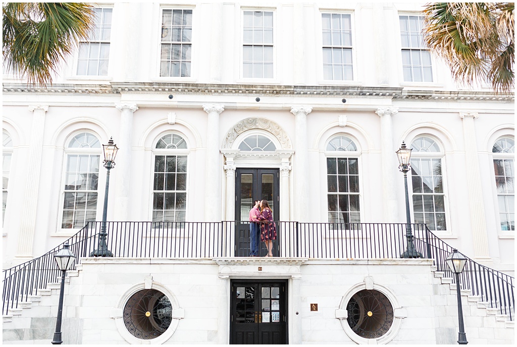 Downtown Charleston engagement session standing at the top of stairs at a white building.