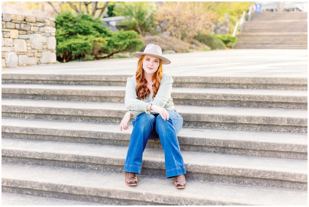High school senior photos of a girl wearing jeans and a green shirt with a tan hat, sitting on steps.