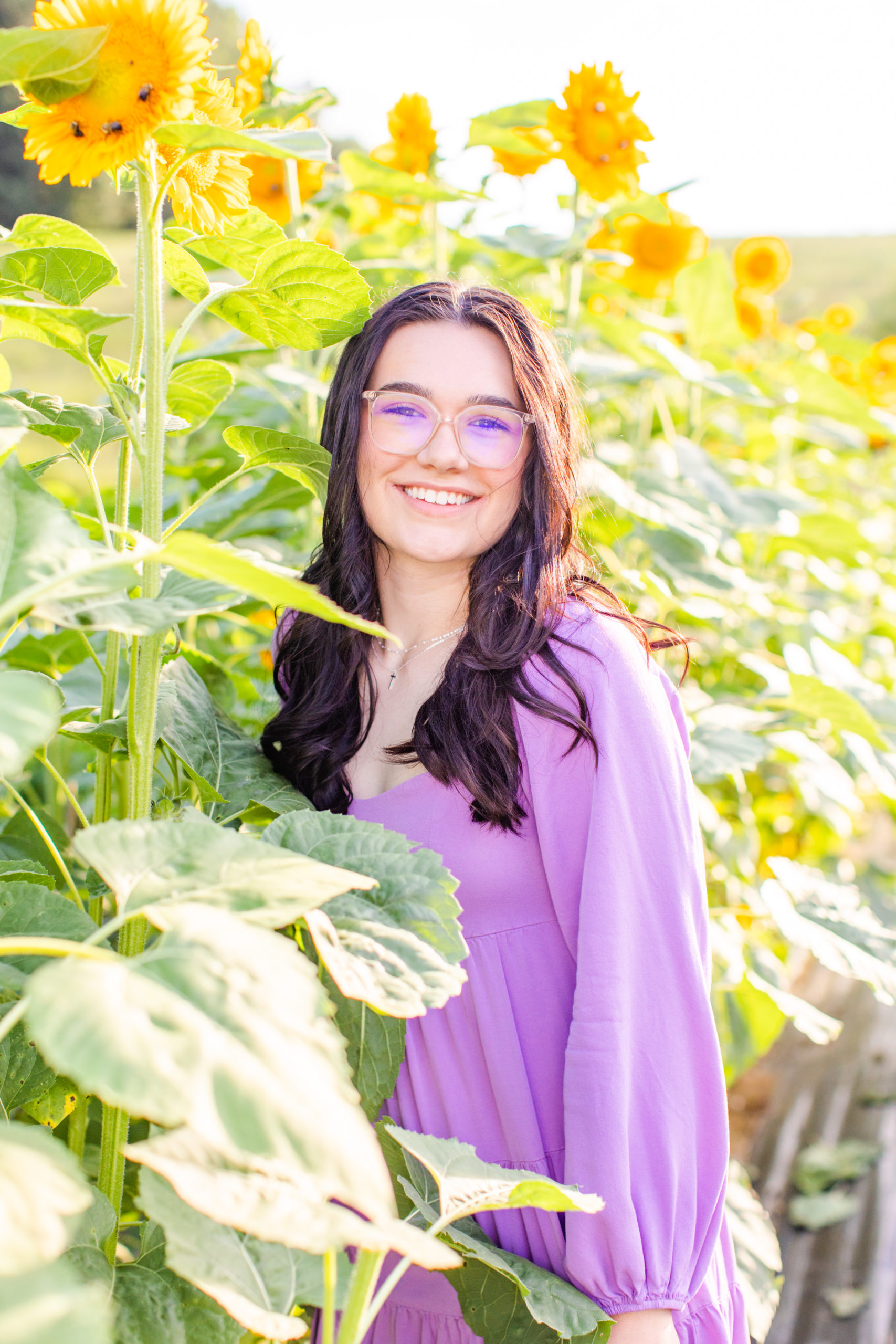 high school senior standing in front of sunflowers in purple dress for senior session at flower farm