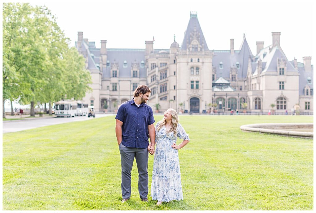 girl in blue long floral dress and guy in navy shirt with grey pants standing in front of biltmore estate holding hands and looking at each other